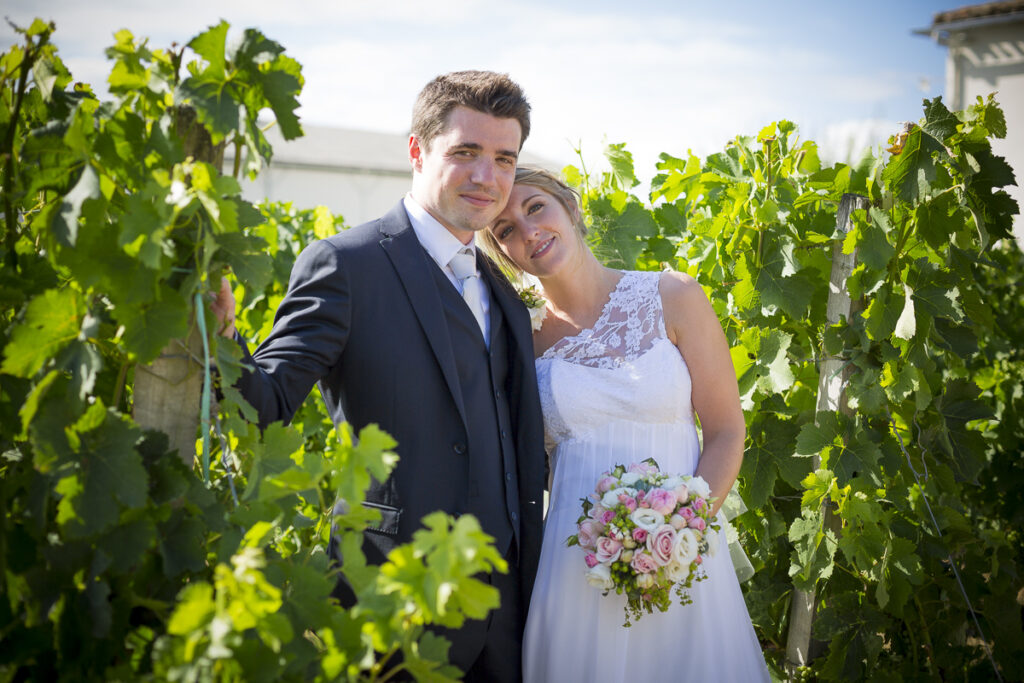 couple de jeunes mariés qui posent entre les vignes au soleil couchant dans les rangs de vignes du chateau Lafitte Laguens à Yvrac dans la région bordelaise, la mariée tient sont bouquet de mariage et pose sa tête sur l'épaule de son mari dans son joli costume bleu de mariage - sebastien huruguen